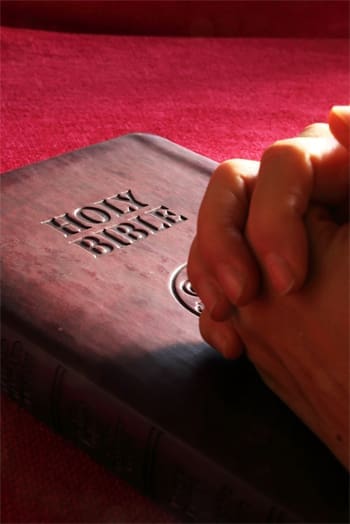 Here's an alt tag for the image: Hands clasped in prayer on Holy Bible.