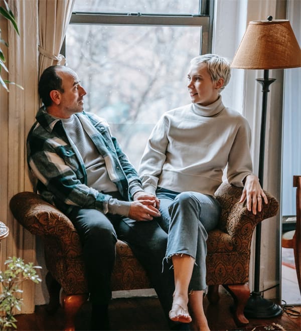 Couple holding hands, seated by window.