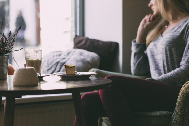 Woman sits alone, coffee, cake, window.